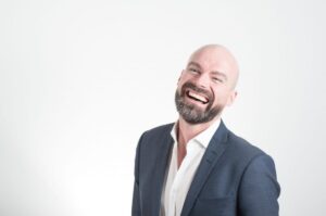 A bald and bearded man smiling widely and showing his straight teeth.